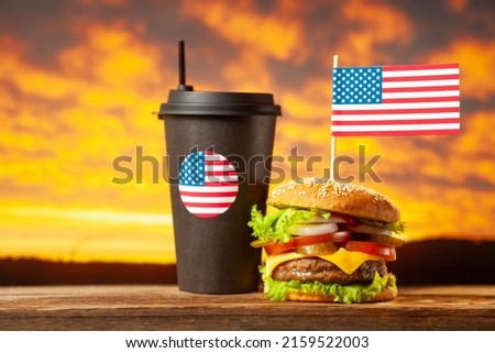 Close-up home made beef burger with american flag on the top and black paper cup with straw on wooden table over sunset background.