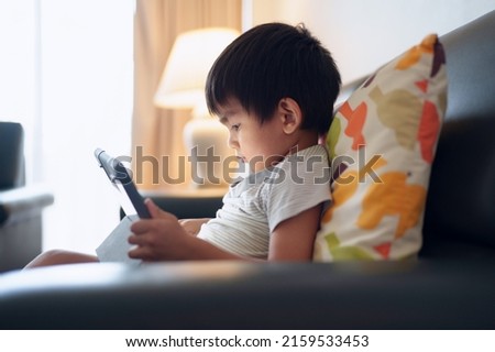 Asian little boy sitting alone on sofa and looking the screen of tablet  in dark room, concept of kid and technology and internet impact for life skill and physical development, online education.