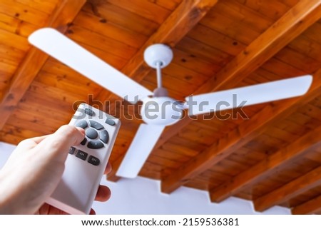A man uses a remote control to turn on a white ceiling fan mounted in a house with wooden ceilings.	 Royalty-Free Stock Photo #2159536381