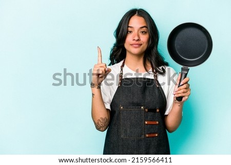 Young hispanic cooker woman holding frying pan isolated on blue background showing number one with finger.