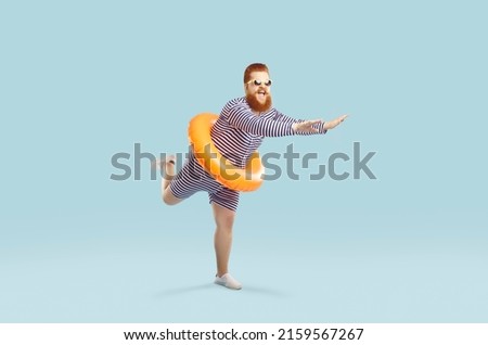 Funny crazy man with inflatable circle pretends to float on light azure background. Cheerful redhead bearded chubby guy in striped leotard having fun with swimming circle at waist. Full length. Banner Royalty-Free Stock Photo #2159567267