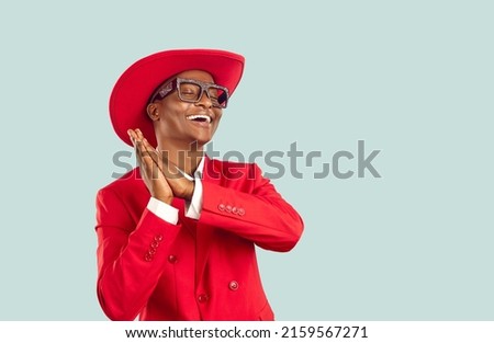 Funny African man in trendy outfit dancing in studio. Happy young Tanzanian guy wearing funky red suit, hat and disco party glasses dancing isolated on blue copy space background. Fashion concept