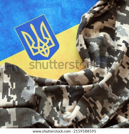 Ukrainian flag and coat of arms with fabric with texture of pixeled camouflage. Cloth with camo pattern in grey, brown and green pixel shapes with Ukrainian trident sign close up.