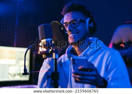 Male sound engineer, podcaster with curly hair, headphones using smartphone in sound studio recording new melody or album.