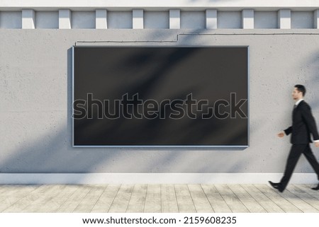 Attractive young european businessman walking past empty black poster hanging on concrete outdoor wall with shadows and sunlight. Billboard, commercial and advertisement concept