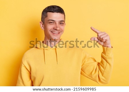 Optimistic positive man wearing casual style hoodie, posing isolated over yellow background, showing a little bit gesture, inch or centimeter, disappointed with minimum size.
