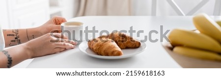 partial view of tattooed woman with coffee cup near croissants and blurred bananas, banner