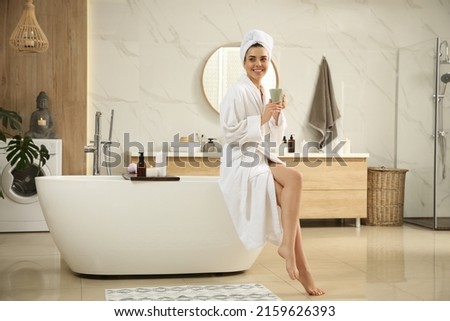 Beautiful young woman with cup sitting on edge of tub in bathroom