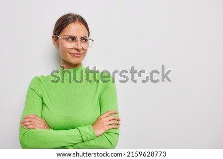 Dissatisfied brunette young woman has sulking expression keeps arms folded feels discontent focused away wears transparent eyeglasses and casual green turtleneck isolated over white background