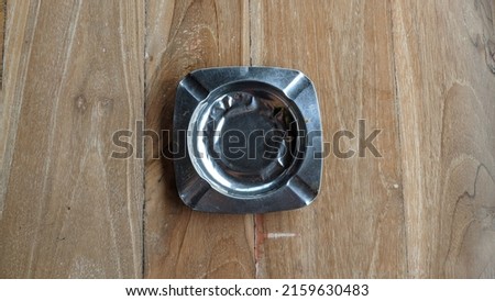 An empty cigarette ashtray in the form of a square box made of iron, is on a wooden table.  Taken from the top corner