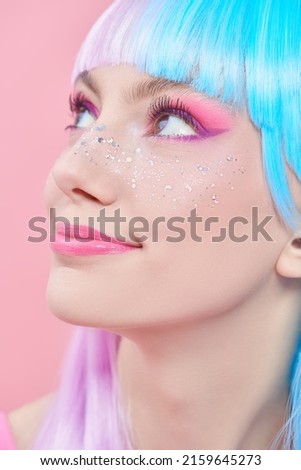 Anime makeup. Pretty girl with bright makeup, glitter freckles and in colored violet-blue wig cutely smiling on a pink background. Hairstyle, hair coloring, make-up. Japanese anime style. 