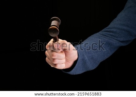 A hand holding either an auctioneers gavel or a judges gavel, like a small hammer, in the right hand. The model is wearing a blue jumper and is photographed with a black background.