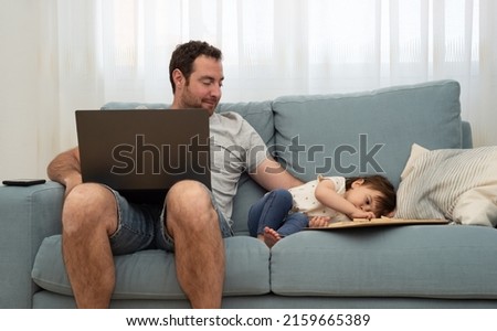 Smiling father sitting on a sofa and looking her little daughter playing close to him during a short break from teleworking.