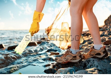 Person collects rubbish in a plastic bag. In the background the sea. Close up of woman's legs. Low angle view. The concept of coastal zone cleaning. Royalty-Free Stock Photo #2159674199