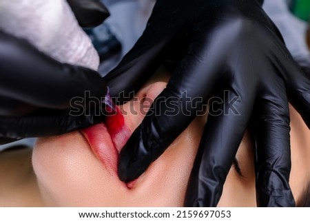 a permanent makeup artist in black gloves holds the corners of the model's lips and stretches them to get a tattoo Royalty-Free Stock Photo #2159697053