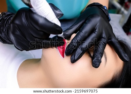 a permanent makeup artist sits in front of a model and does lip tattooing with a tattoo machine and red lip pigment Royalty-Free Stock Photo #2159697075
