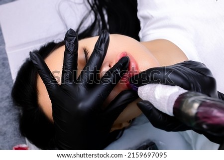 a permanent makeup artist holds a tattoo machine in his hand and applies permanent makeup to the lips of a model with red pigment Royalty-Free Stock Photo #2159697095