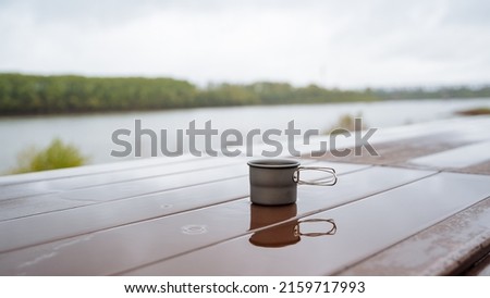 Coffee mug in nature, camping utensils, drinking hot tea on a hike, reflecting the silhouette of a glass in a puddle of water, a cup standing on the table wet, rain in the forest. High quality photo