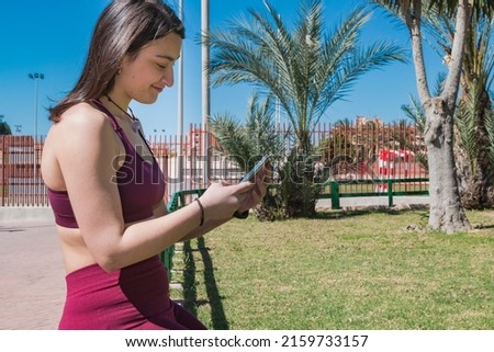 A young brown-haired Caucasian girl dressed in sportswear is sitting on a green wooden fence in a public park while looking at her cell phone and touching its screen.