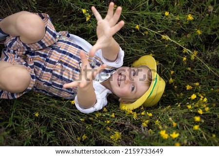 Portrait of a cute cheerful boy in a yellow hat, lying on a field of wild yellow flowers. Summer mood, carefree joyful childhood, outdoor recreation. Hello summer, energy of nature. view from above