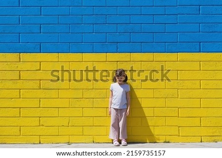 little toddler girl sad on the background of a brick wall painted in the colors of the Ukrainian flag, the concept of supporting the children of Ukraine during the war.