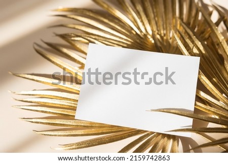 Invitation card mockup with golden palm leaves on beige pastel background. Top view, flat lay, copy space. Template blank of white paper mock up for branding and advertising