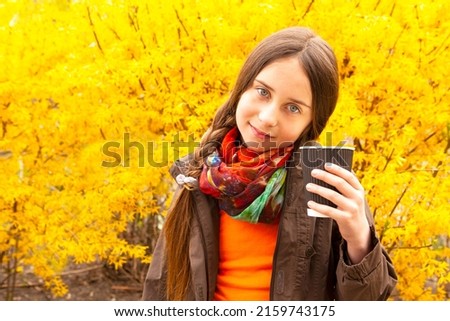A cute brunette girl holds a paper cup with hot coffee, tea against the background of yellow flowering bushes. A beautiful girl enjoys a fragrant cappuccino.