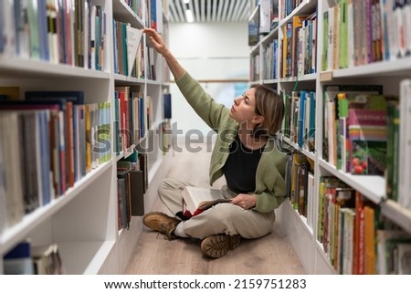 Thoughtful Scandinavian middle-aged pretty woman student getting second higher education, preparing for test exams in university library, leaning towards shelf with books, holding a textbook in hands.