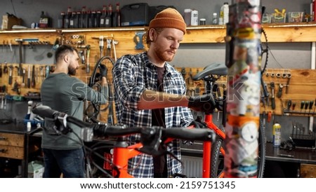 Cycling mechanic fixing bicycle seat while his male colleague checking bicycle wheel spoke with bike spoke key on background in workshop. Young caucasian repairmen. Bike service, repair and upgrade Royalty-Free Stock Photo #2159751345