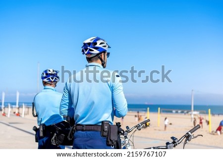 Two policeman patrolling seaside promenade on bicycles. People are sunburning on the city public beach on the Atlantic shore. Bike police patrol on the ocean beach Royalty-Free Stock Photo #2159767107