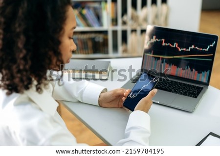 Confident smart dark-skinned girl, trader, stock investor, sits in office, uses smart phone and a laptop, works with quotes charts, analyzes the dynamics of the cost of crypto currency Royalty-Free Stock Photo #2159784195