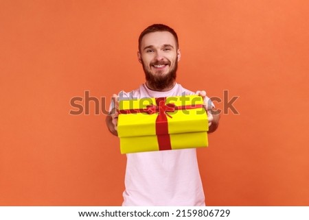Portrait of happy bearded man giving gift box to camera with excited smile, greeting on holiday and sharing present, wearing pink T-shirt. Indoor studio shot isolated on orange background.