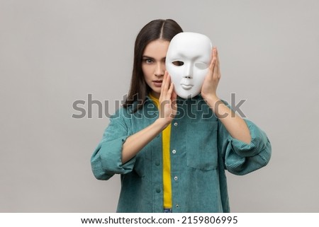 Portrait of strict bossy woman covering half of face with white mask, multiple personality disorder, wearing casual style jacket. Indoor studio shot isolated on gray background. Royalty-Free Stock Photo #2159806995