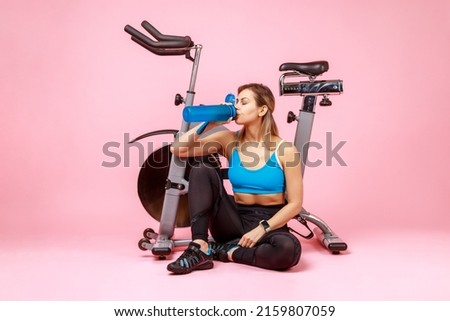 Portrait of thirsty athletic woman drinking water from bottle, restoring water balance after exhausted workout, wearing sports tights and top. Indoor studio shot isolated on pink background.