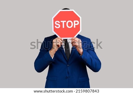 Portrait of man hiding face behind Stop symbol, red traffic sign warning of restricted access, banned service, wearing official style suit. Indoor studio shot isolated on gray background.