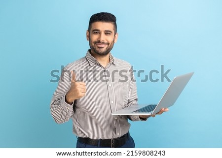Portrait of smiling positive businessman standing with portable computer in hand, looking at display, showing thumb up, wearing striped shirt. Indoor studio shot isolated on blue background. Royalty-Free Stock Photo #2159808243