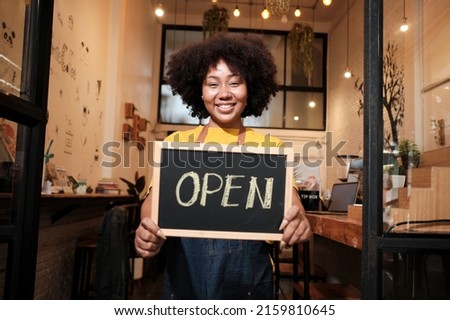 One African American female startup barista stands at casual cafe door, looks at camera, and shows open sign, happy and cheerful smiles with coffee shop service jobs, and new business entrepreneurs.