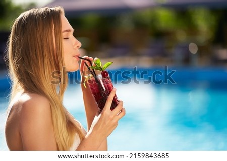 Pretty woman drinks refreshing cocktail decorated with mint, cherry and lemon. Female enjoys a drink near swimming pool with blue water. Girl chilling with beverage in tropical sun. Vacation concept Royalty-Free Stock Photo #2159843685