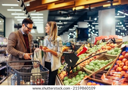 Cheerful couple choosing fruit at groceries department of the supermarket. Man holding mesh bag and woman holding fruits. Shopping at the modern mall Royalty-Free Stock Photo #2159860109