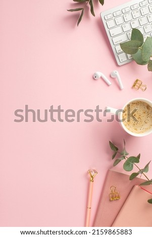 Business concept. Top view vertical photo of workspace keyboard wireless earbuds cup of coffee girlish pens pink notepads gold binder clips and eucalyptus on isolated pink background with copyspace