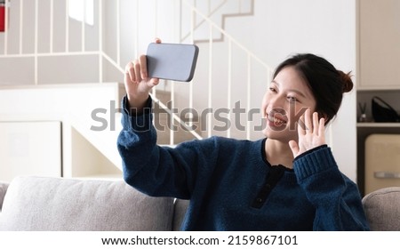 Smiling young asian woman using mobile phone while sitting on a couch at home with laptop computer
