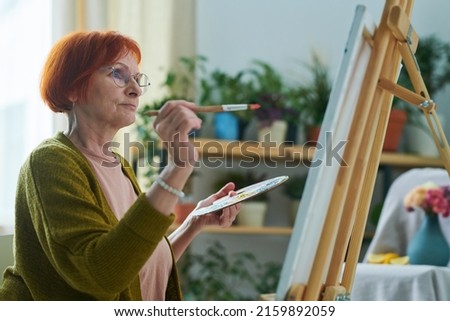 Senior redhead woman in eyeglasses sitting in front of easel and using palette and paintbrush to paint a picture as art therapy
