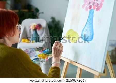 Red haired senior woman in eyeglasses sitting in front of easel and painting still life with palette