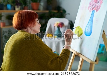 Redhead mature woman in eyeglasses learning to paint still life with paintbrush on easel