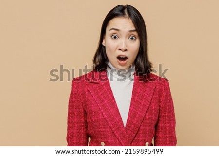 Shocked surprised astonished scared woman of Asian ethnicity wear red jacket look camera with open mouth isolated on plain pastel beige background studio portrait. People lifestyle fashion concept.