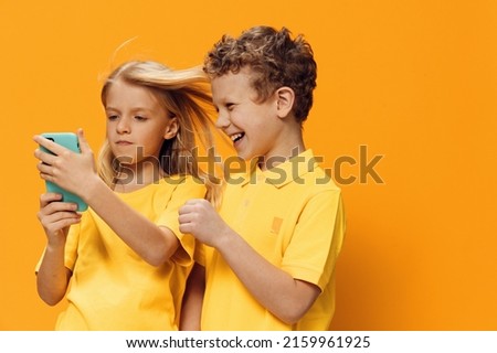 cute, funny children, brother and sister of school age are standing in bright clothes on a yellow background and the girl is holding a phone in a blue case in her hands