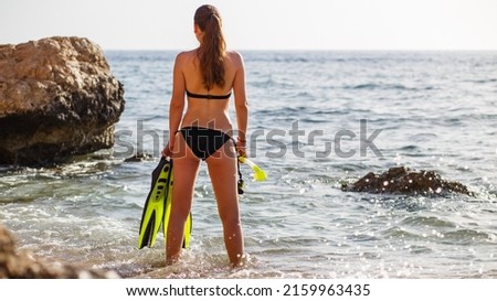 Young attractive woman in bikini holding flippers standing in water in the morning.