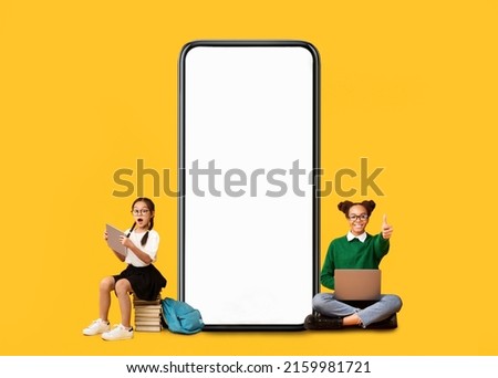 Wow, Great Offer. Excited Multiethnic School Girls Sitting On Floor And Pile Stack Of Books Near Big Giant Cell Screen, Using Pc Laptop And Tablet On Yellow Orange Studio Background, Showing Thumbs Up