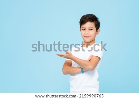 Cute smiling boy in plain white t shirt opening empty hand in isolated studio light blue color background Royalty-Free Stock Photo #2159990765