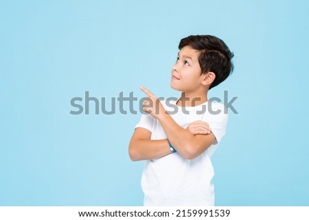 Cute smiling boy in plain white t shirt looking and pointing hand up in isolated studio light blue color background